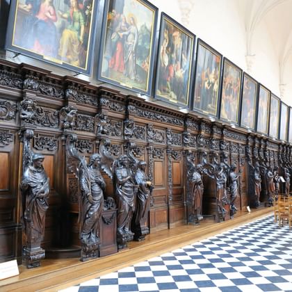 Ornate wooden choir stalls with carved figures in St. Paul's Church, Antwerp. Black and white checkered floor, paintings above, visitors walking.