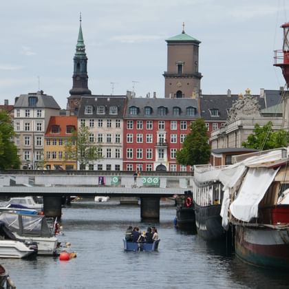 Copenhagen harbor with boats, colorful historic buildings, church spires, and a red lighthouse. A bridge crosses the canal in the foreground.