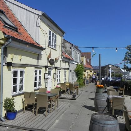 Bunte historische Gebäude mit Außenbestuhlung entlang der alten Hafenpromenade in Køge, Dänemark. Gelbe Fassaden mit roten Ziegeldächern unter blauem Himmel.