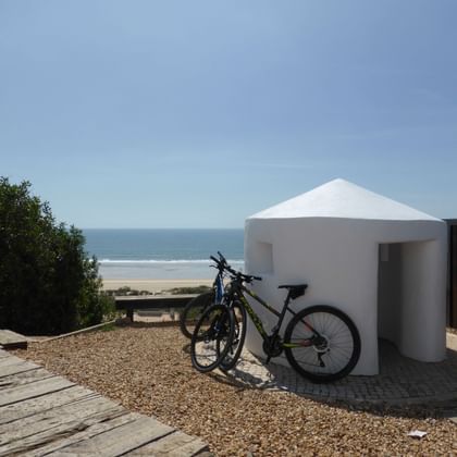 Zwei Fahrräder neben einem weißen zylindrischen Strandunterstand auf Kiesboden, mit Meer und Sandstrand im Hintergrund.