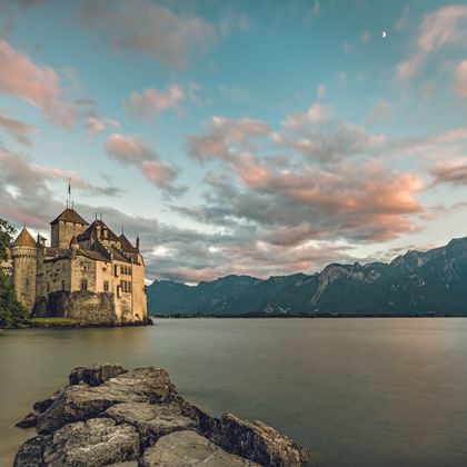 Chillon Castle on Lake Geneva with mountains in background. Rocky shore in foreground, dramatic sunset sky with pink clouds and crescent moon visible.