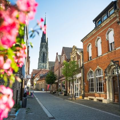 Kopfsteinpflasterstraße in Billerbeck mit roten Backsteingebäuden, rosa blühendem Baum im Vordergrund und gotischem Kirchturm im Hintergrund.