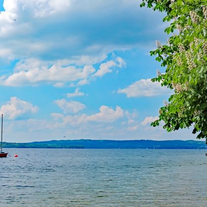 Scenic view of Lake Starnberg with a red sailboat anchored on calm blue water. Green tree branches frame the top right, blue sky with white clouds above.