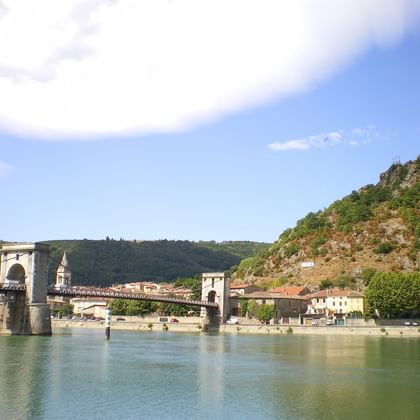 Historic stone bridge with towers crossing the Rhône River, with a hillside village and green cliffs under blue sky near Lyon.