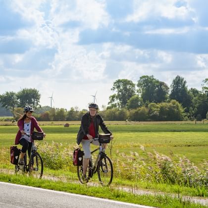 Two cyclists with touring bikes and panniers stopped on a country road beside green fields. Wind turbines visible in the distance under cloudy sky.