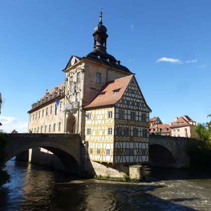 Bambergs Altes Rathaus auf einer Steinbrücke über dem Fluss mit barocken Fresken und Fachwerkarchitektur unter blauem Himmel.