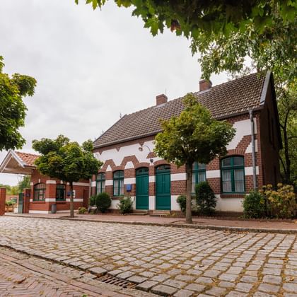 Historisches Backsteingebäude mit grünen Fensterläden und Ziegeldach des Textilmuseums Bocholt, umgeben von Kopfsteinpflaster und Bäumen.