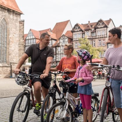 Family of four with bicycles on cobblestone market square in Hann. Münden, with historic half-timbered buildings and stone church in background.