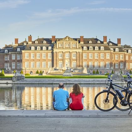 Zwei Radfahrer sitzen am Wasser und betrachten Schloss Nordkirchen, ein barockes Schloss mit formalen Gärten. Ihre Fahrräder stehen daneben.