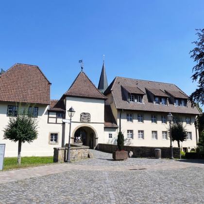 White castle complex in Bersenbrück with red tile roofs, arched entrance gate, and twin church spires under blue sky.