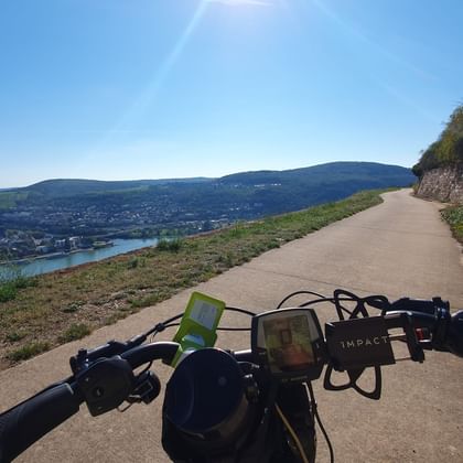 Blick vom E-Bike-Lenker auf befestigten Weg entlang Fluss mit Stadt und Hügeln im Hintergrund. Steinmauer rechts, grüne Hänge links unter blauem Himmel.