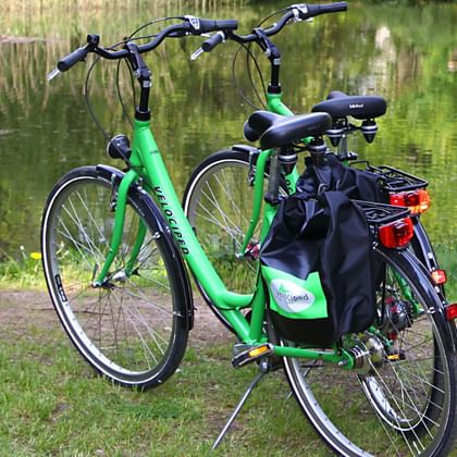 Grünes Velociped 7-Gang Tourenrad mit schwarzen Packtaschen auf Gras am See geparkt, mit weißer Bank im Hintergrund sichtbar.