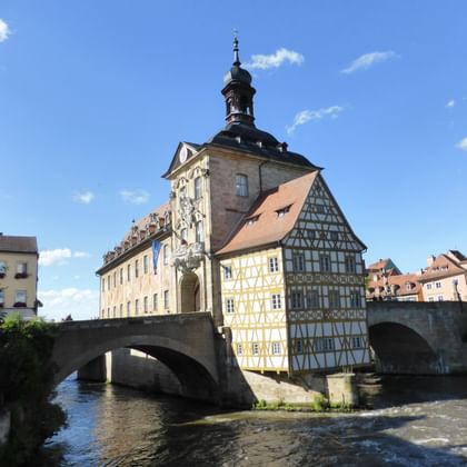 Historic Bamberg city gate with baroque tower and half-timbered buildings on stone bridge over flowing river under blue sky with clouds.