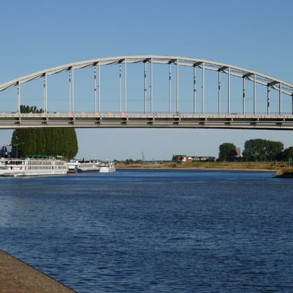 White arched John Frost Bridge crossing a wide river in Arnhem with passenger boats below and green trees lining the banks under clear sky.