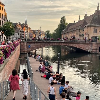 People sitting along a canal waterfront in Strasbourg with a stone bridge, historic buildings, and flower-lined quay under an evening sky.