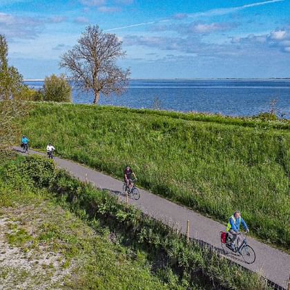 Aerial view of cyclists on paved path through green coastal landscape near Muiden, Holland. Blue water and trees visible in background.
