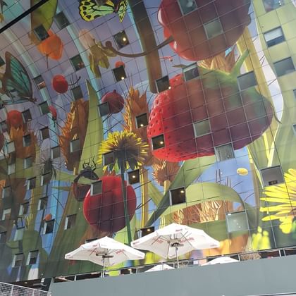 Vibrant ceiling mural in Rotterdam market hall featuring oversized fruits, vegetables, and flowers in bold colors reflected on glass facade.