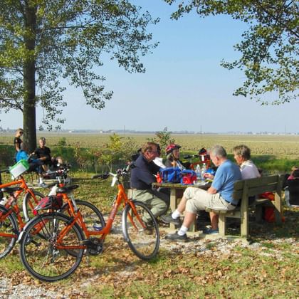 Gruppe von Radfahrern rastet an Holzbänken unter Bäumen. Orange Fahrräder stehen daneben, flache Felder im Hintergrund.