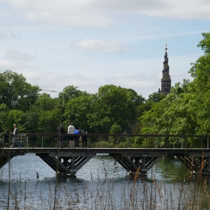 Cyclists and pedestrians on Dyssebroen bridge crossing water in Copenhagen. Church spire visible behind green trees under cloudy sky.