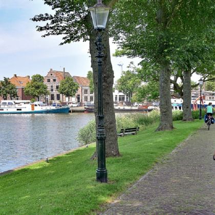 Cyclist on paved path beside harbor in Medemblik with boats, historic Dutch buildings, and green waterfront park under cloudy sky.