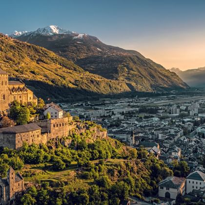 Historic castle on a hill overlooking Sion in Valais at sunset, with snow-capped mountains and the Rhone valley in the background.