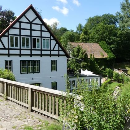 White half-timbered building with black beams beside a stream in Aumühle. A wooden footbridge crosses the water, surrounded by lush greenery.
