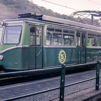 Green and white Drachenfels Railway tram with golden crest logo at a station platform. Hills and autumn trees visible in background.