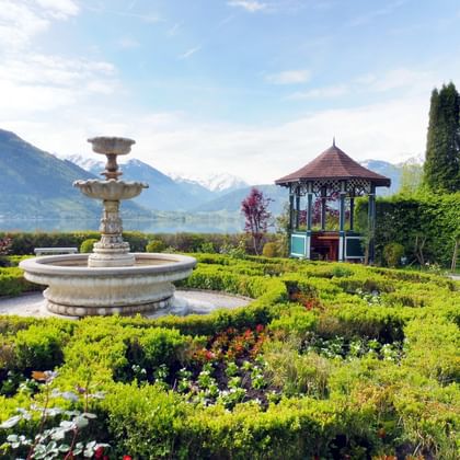 Ornate fountain and gazebo in manicured garden at Zell am See with lake and snow-capped Alps in background under blue sky with clouds.