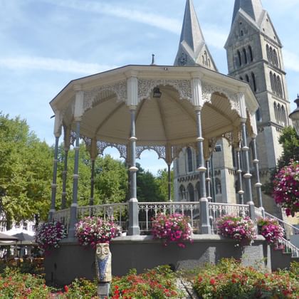 White ornate gazebo with decorative columns and pink flower baskets in foreground, with twin-towered Munsterkerk Catholic Church visible behind.