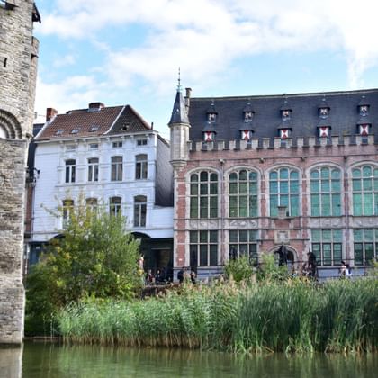 Mittelalterlicher Steinturm und verziertes Backsteingebäude mit großen Bogenfenstern spiegeln sich im Kanalwasser, mit Schilf im Vordergrund in Gent.