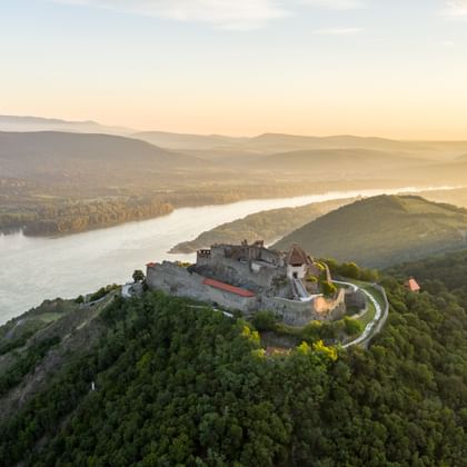 Luftaufnahme der Burg Visegrad auf einem bewaldeten Hügel mit Blick auf die Donaukurve bei Sonnenuntergang im goldenen Licht.
