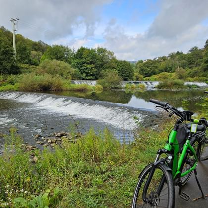 Two green bicycles with panniers parked on paved path beside Lahn River with cascading weir and lush green vegetation under cloudy sky.