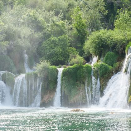 Mehrere kaskadierende Wasserfälle im Krka-Nationalpark fließen über moosbewachsene Kalksteinterrassen in türkisfarbene Becken, umgeben von üppigem Wald.