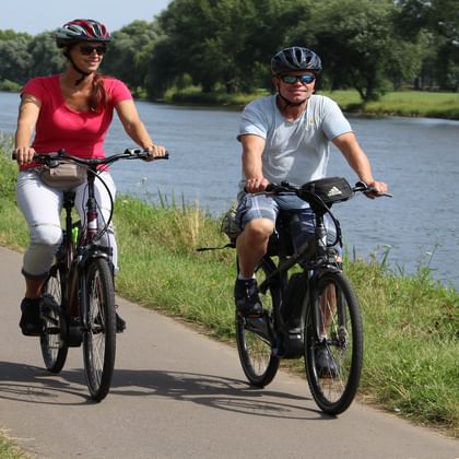 Two cyclists with helmets riding e-bikes on paved path along Weser River. Woman in red shirt and man in white shirt cycling beside water.