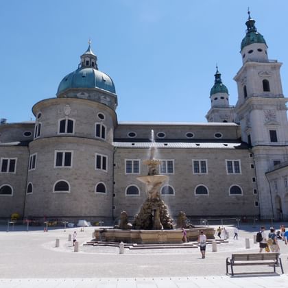 Salzburger Dom mit grüner Kupferkuppel und zwei Glockentürmen. Barocker Brunnen mit Skulpturen steht auf dem Platz mit Besuchern.