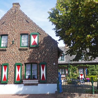 Traditional Dutch brick house along Rurufer cycle path with red and white decorative shutters, green window frames, and a large tree.