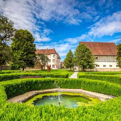 Historisches Wasserschloss Glatt mit Fachwerkgebäude und Brunnen in gepflegtem Garten mit Heckenanlage unter blauem Himmel bei Sulz am Neckar.