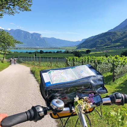 Fahrradlenker mit Kartenhalter auf Radweg durch Apfelhaine bei Bozen, mit Bergen und See im Hintergrund.