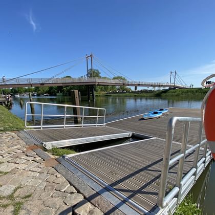 Wooden dock with metal railings and a blue kayak on the Elbe River in Geesthacht. A suspension bridge spans the water in the background.
