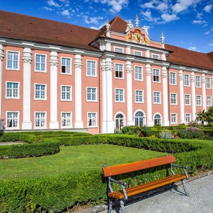 Schloss Meersburg mit Barockarchitektur Schloss Meersburg, ein rosa Barockpalast mit weißen Säulen und verzierter Fassade, umgeben von gepflegten Gärten mit Holzbank im Vordergrund.