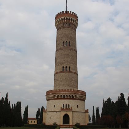 Hoher zylindrischer Steinturm mit Zinnen und Bogeneingängen, umgeben von Zypressen unter bewölktem Himmel in Solferino.