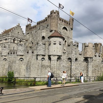 Medieval stone fortress Gravensteen in Ghent with high walls, towers and battlements. Flags fly from the castle, tourists walk nearby.