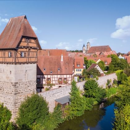 Aerial view of Dinkelsbühl showing a medieval stone tower with timber-framed upper section and red tile roof beside a river with traditional buildings.