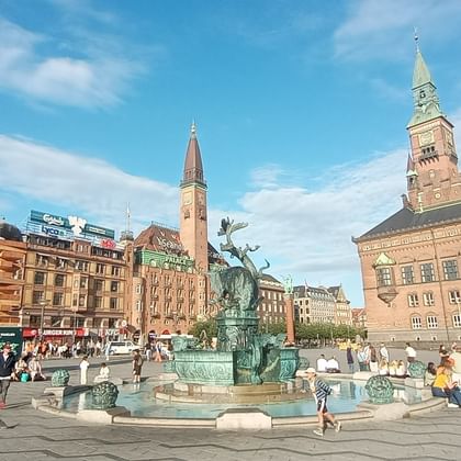 Dragon Fountain at Rådhuspladsen in Copenhagen with historic buildings. Bronze fountain with dragon sculptures in center of square, surrounded by people.