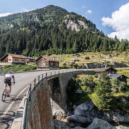 Cyclists on stone bridge in Curaglia-Sedrun Two cyclists crossing a stone arch bridge in Curaglia-Sedrun with traditional Swiss chalets and forested mountains in the background.