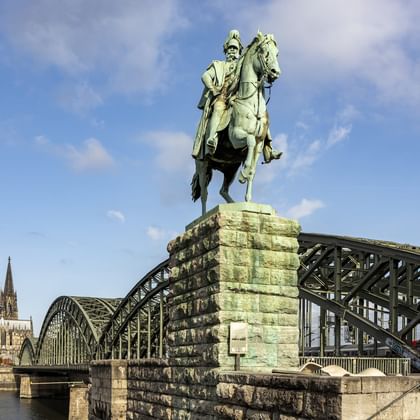 Bronze equestrian statue on stone pedestal at Hohenzollern Bridge with Cologne Cathedral's twin spires visible in background under blue sky.