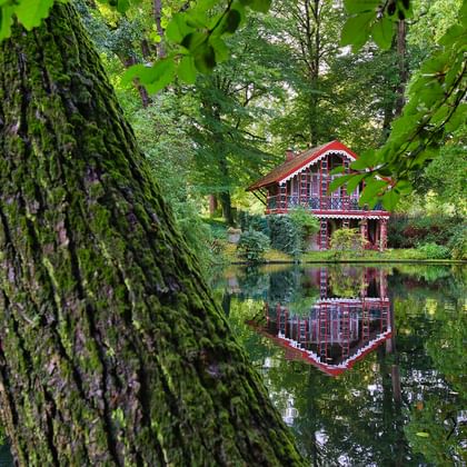Red wooden house with white trim reflected in calm lake water, surrounded by lush green trees and vegetation in Cuxhaven.