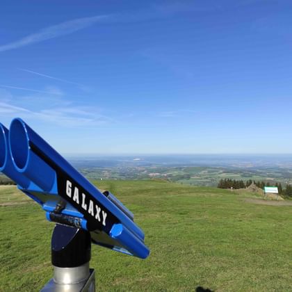 Blaues Galaxy-Fernrohr auf einem Hügel mit weitem Blick über grüne Felder und ferne Ebenen unter klarem blauen Himmel.