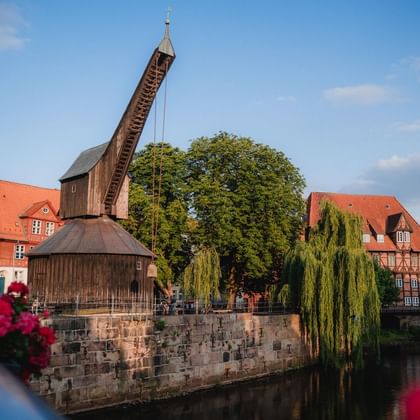 Historischer Holzkran auf Steinfundament am Fluss in Lüneburg. Gebäude mit roten Dächern und Trauerweide im Hintergrund unter blauem Himmel.