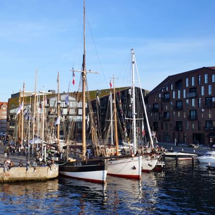 Historic wooden sailing ships moored in Christianshavn canal, Copenhagen. People sit along the waterfront with modern buildings in the background.